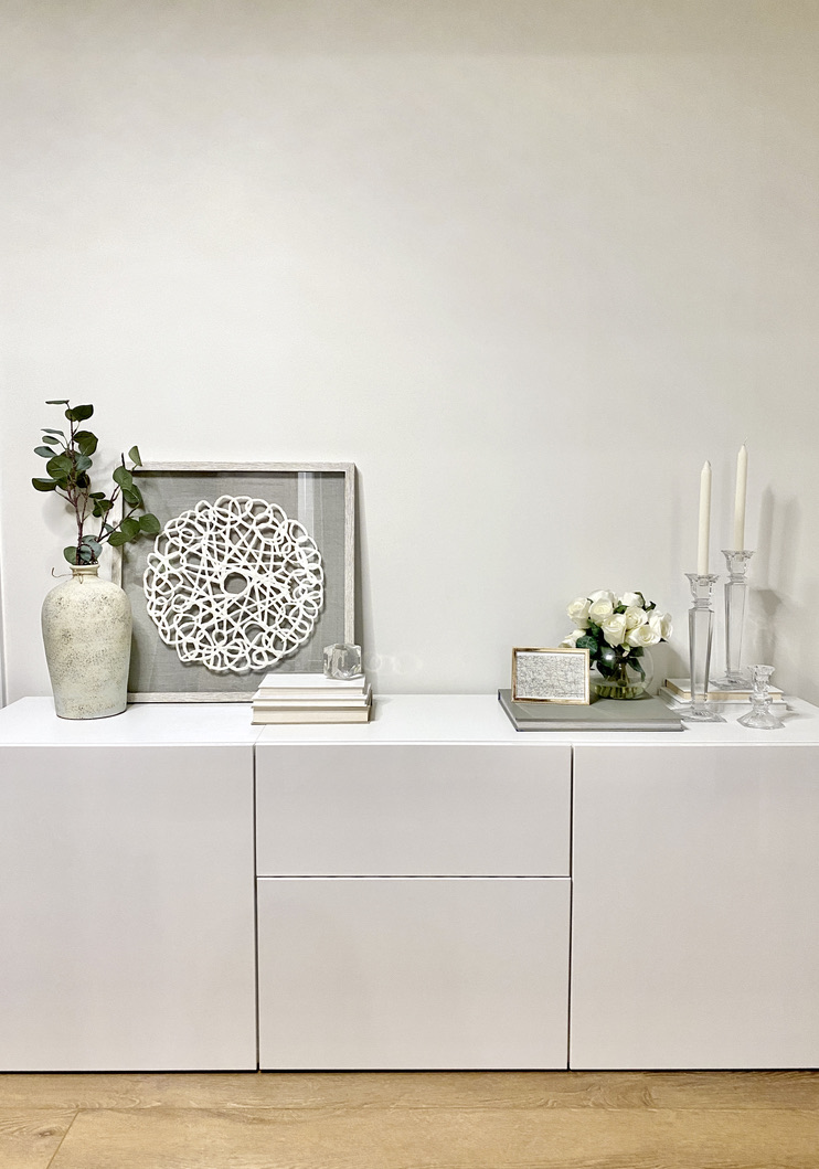 brown LVP wood floor and white besta sideboard styled with soft neutral decor: glass candleholders, hardcover books, leaning paper machier art, beige stone textured vase and greenery, white roses, walls are painted benjamin moore white dove - calgary interior design, bridgeland